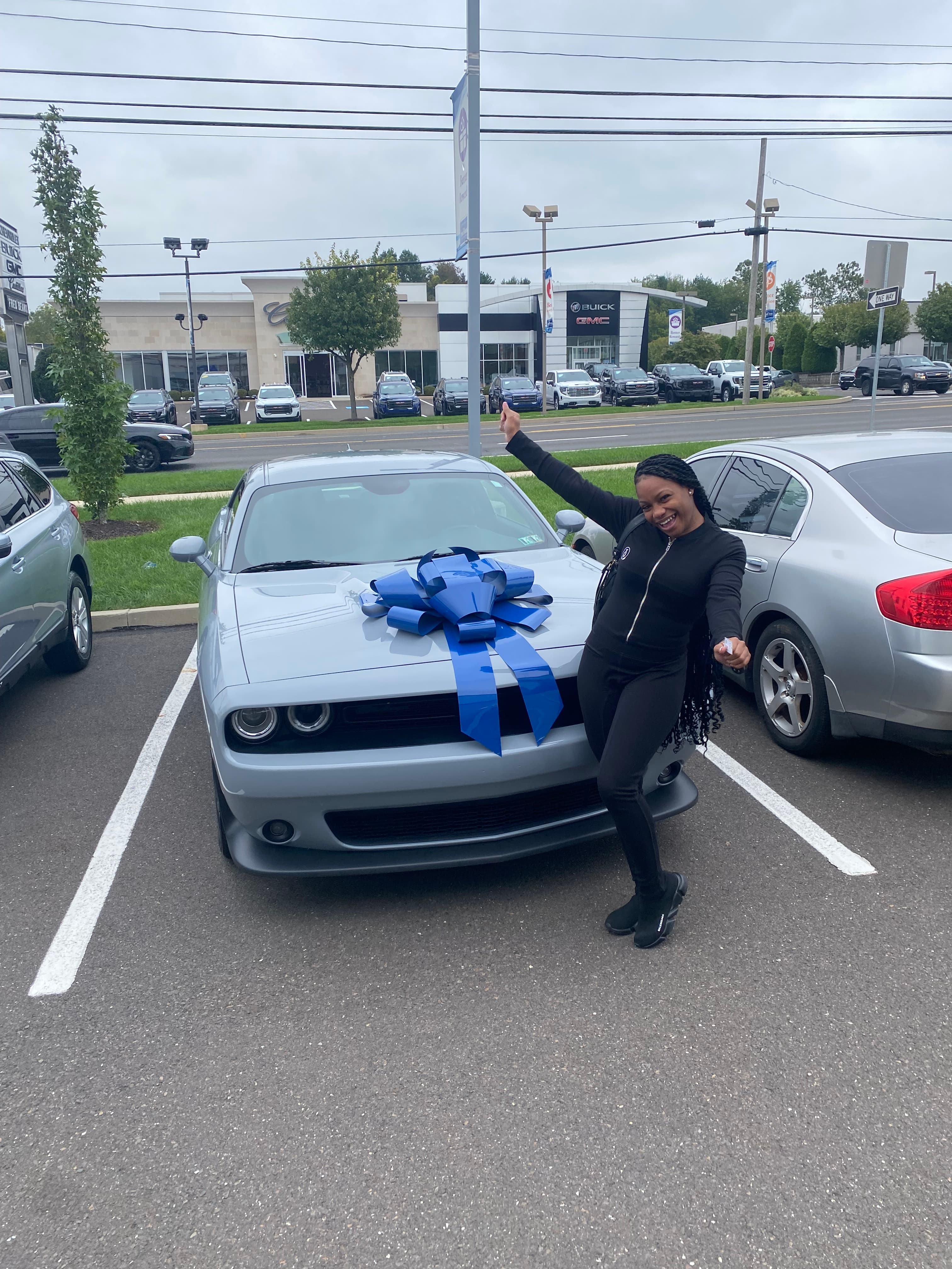 Smiling woman poses with a silver car and a large blue bow at a dealership.