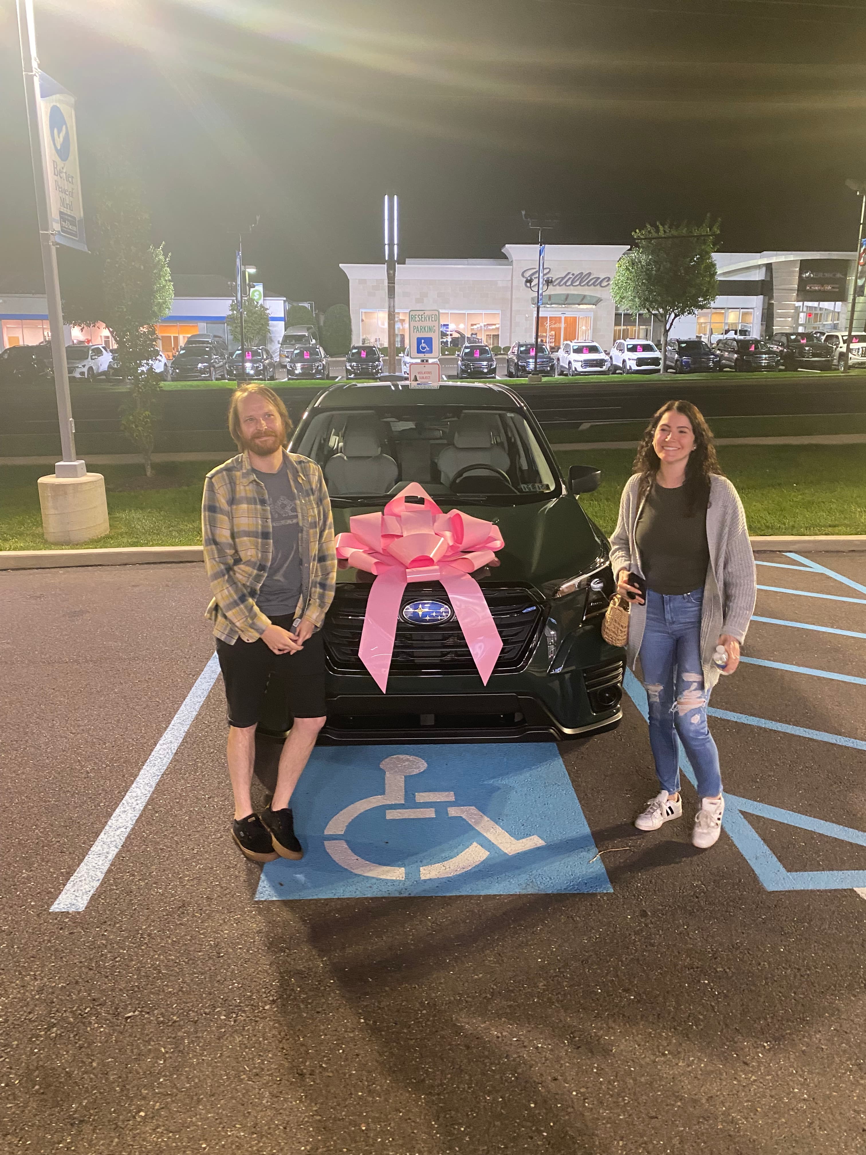 A couple poses with a new dark green Subaru featuring a large pink bow.