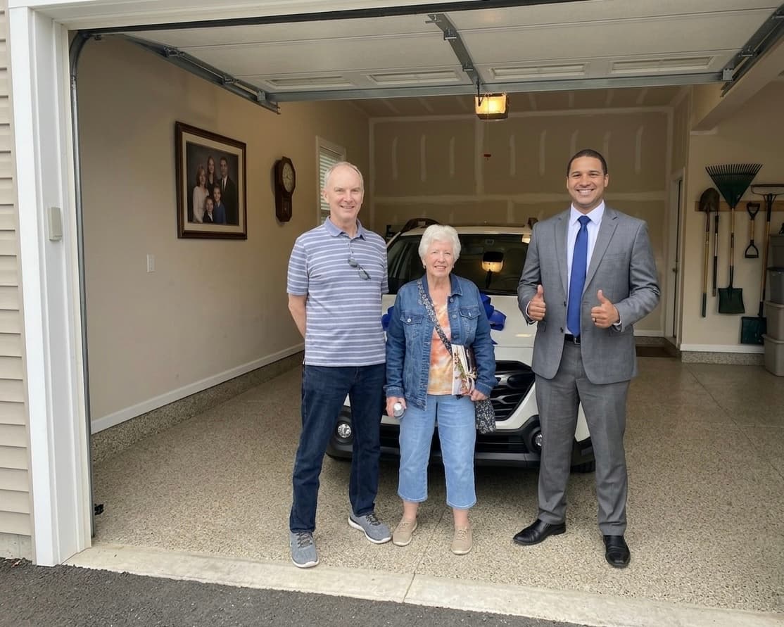 Older couple and salesman pose with a new white car with blue bow in garage.
