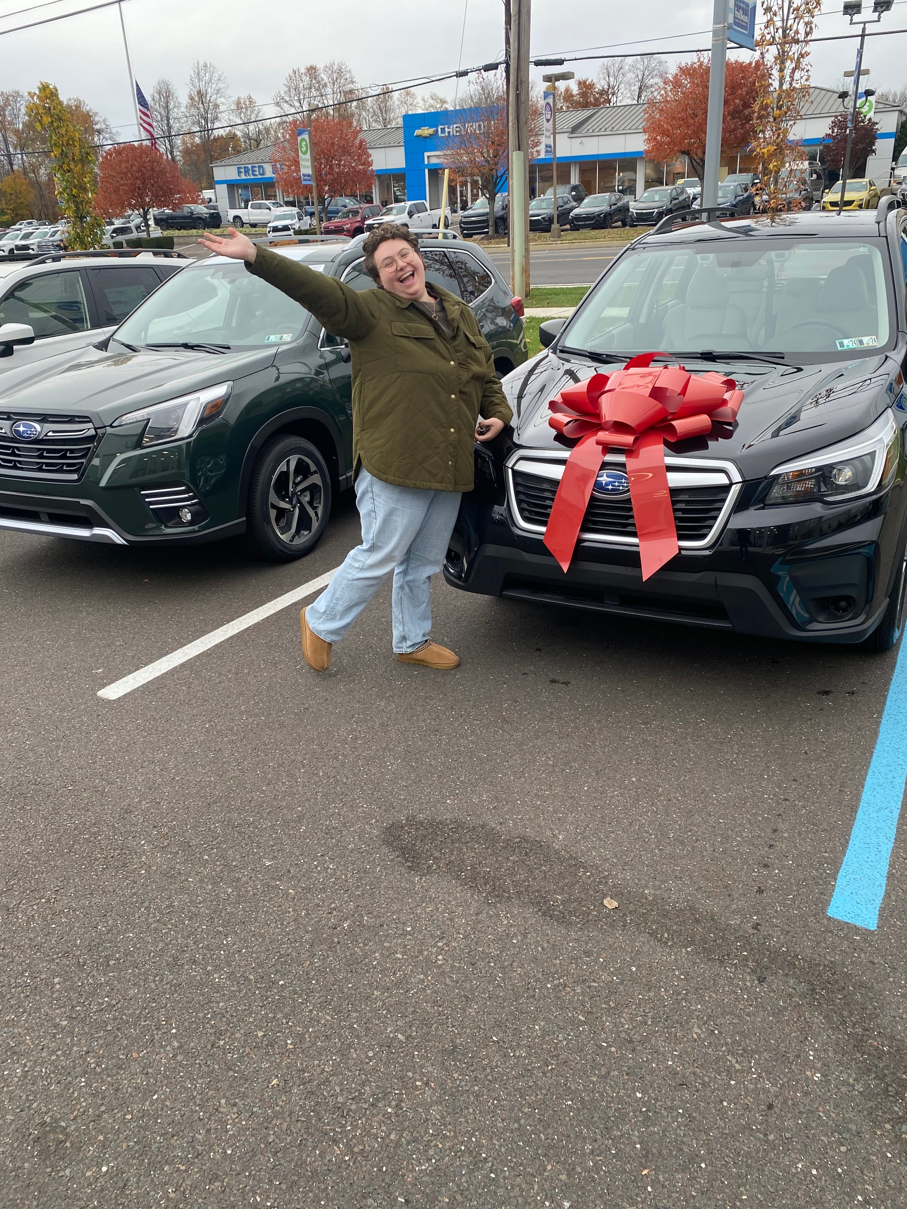 Smiling person in a green jacket stands beside a black car with a red bow.