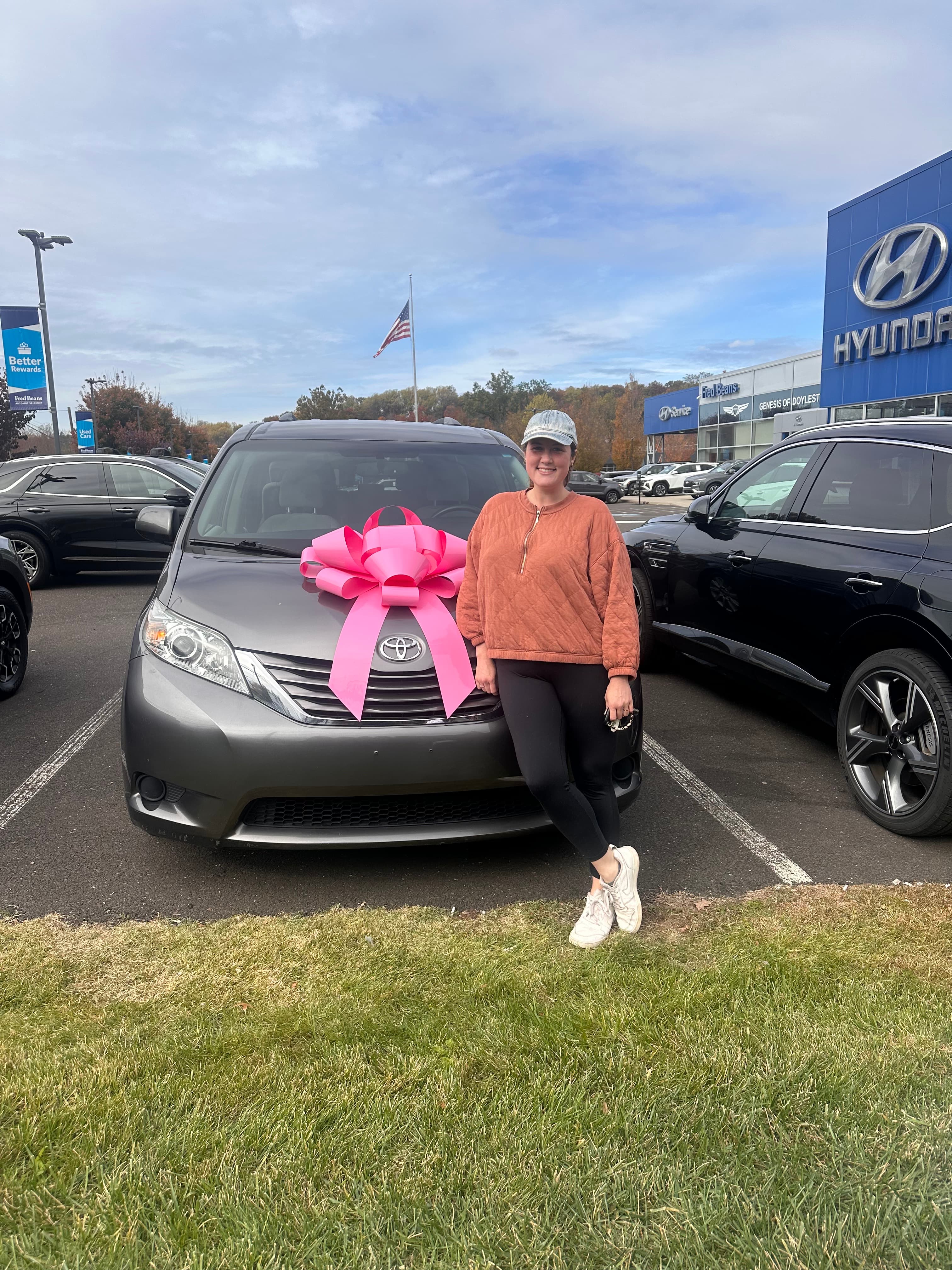 Smiling woman sits on the hood of a grey Toyota with a large pink bow.