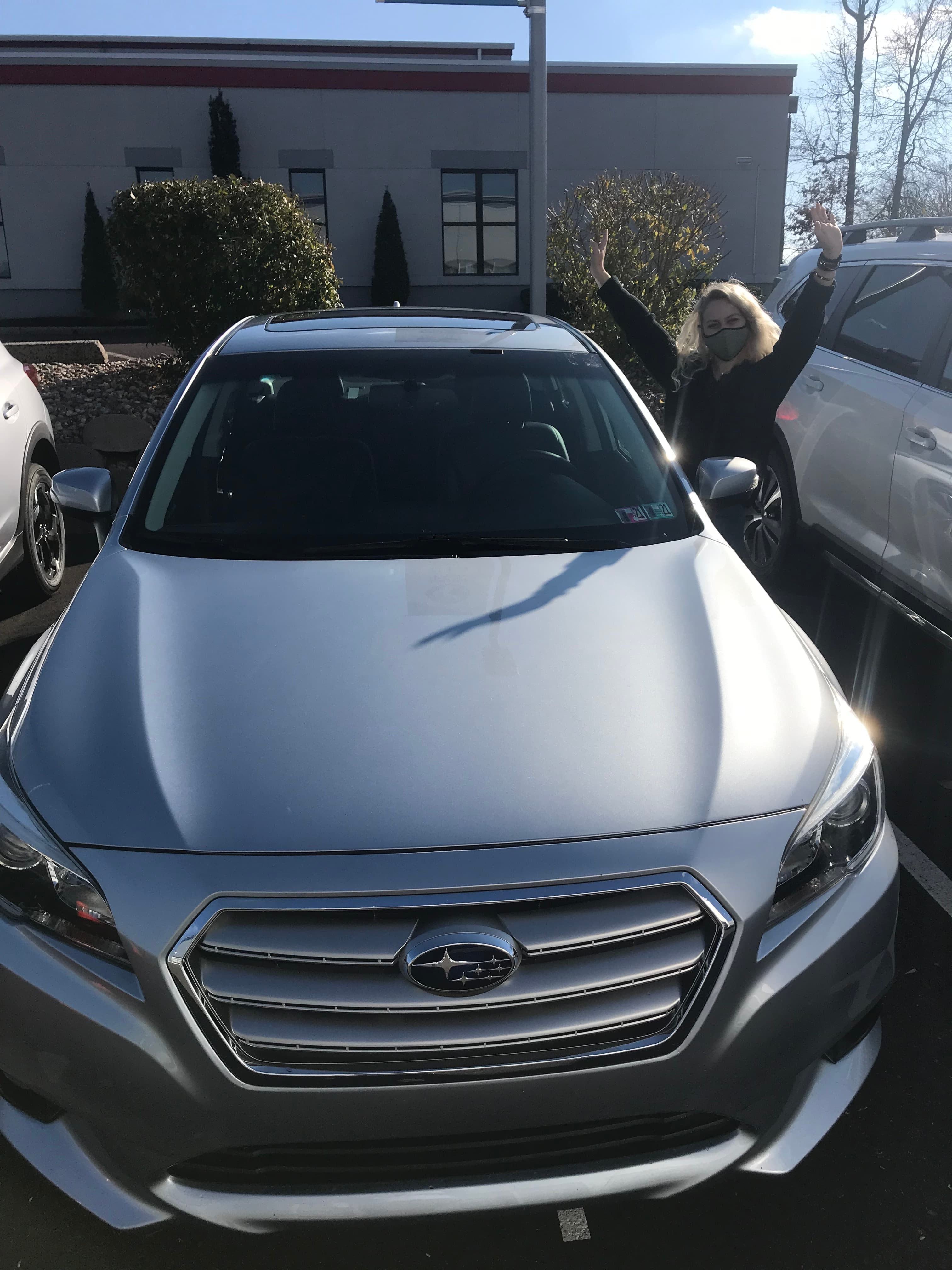 Woman celebrating with arms raised behind a silver Subaru SUV in a parking lot.