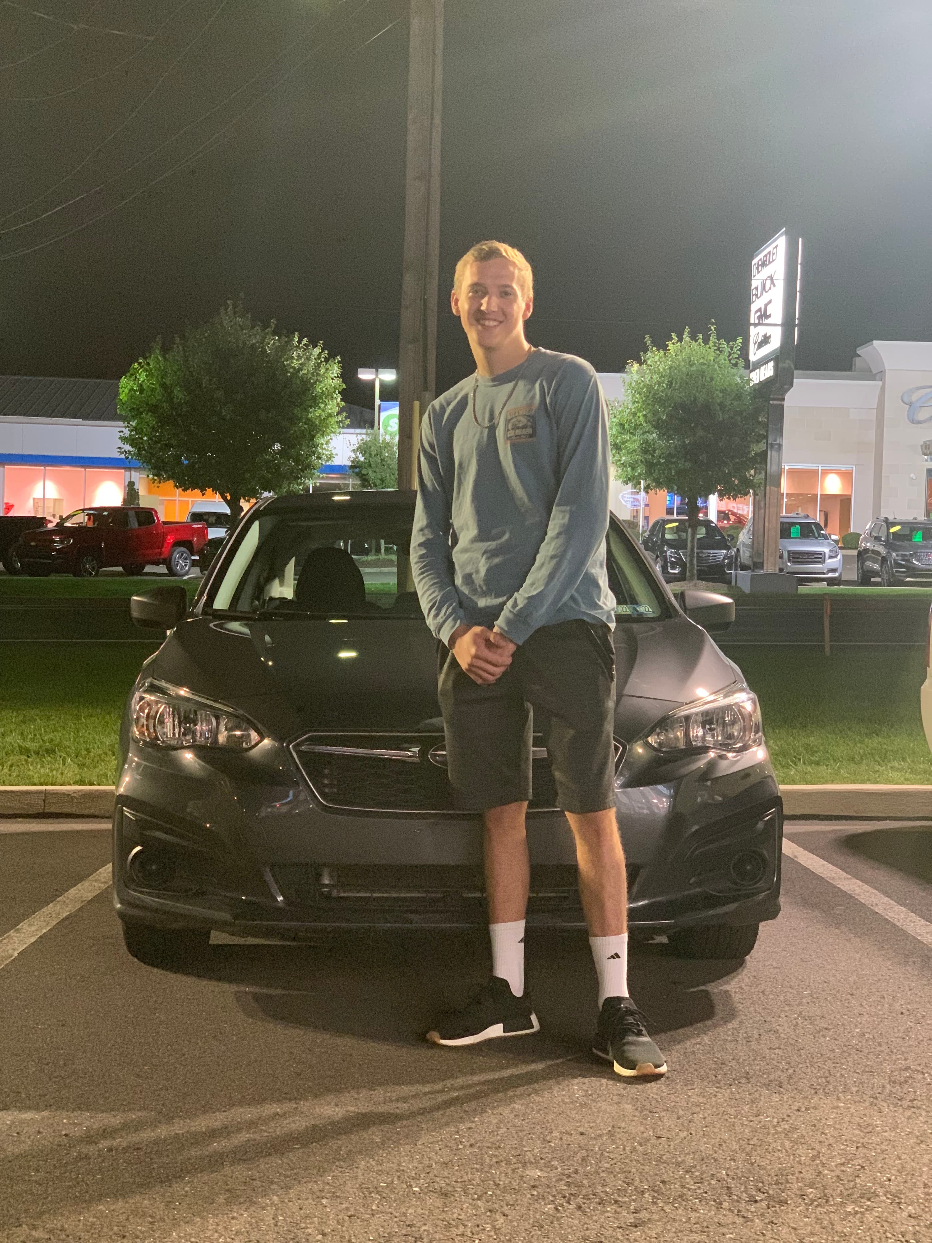 Smiling man in blue shirt stands before a grey car at a dealership at night.