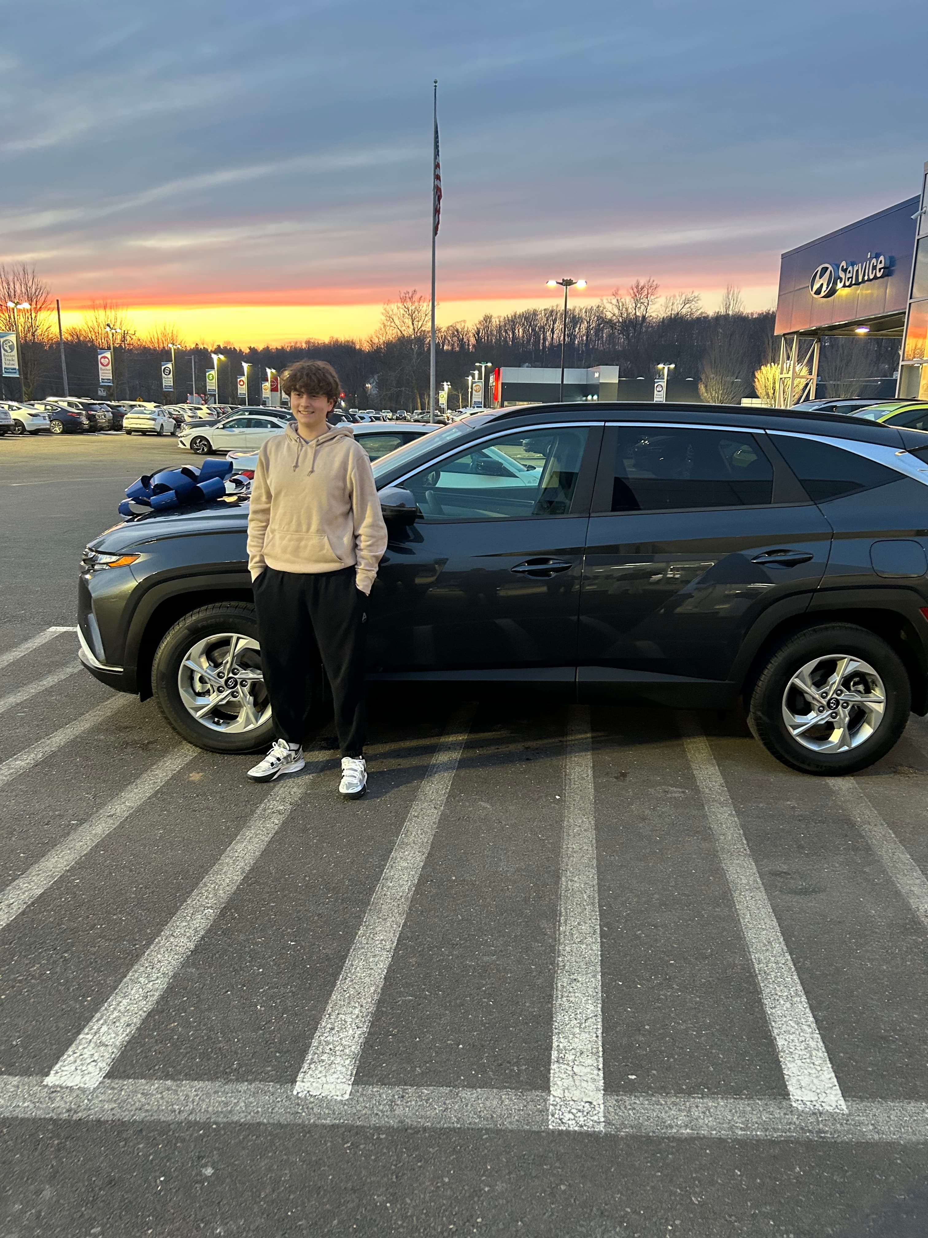Young man posing with a new dark grey SUV and blue bow at sunset.