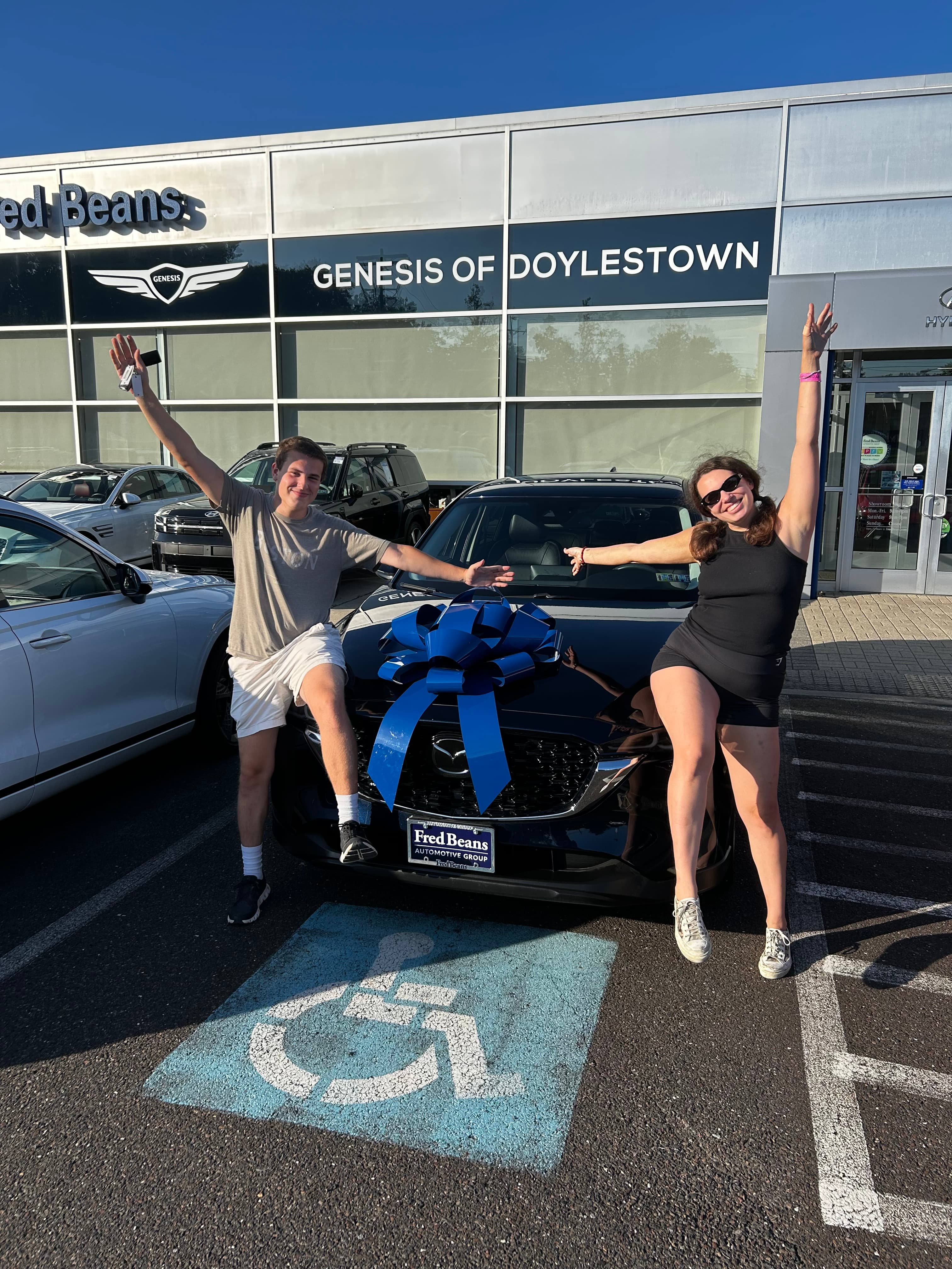 Excited couple poses with a new black car adorned with a large blue gift bow.